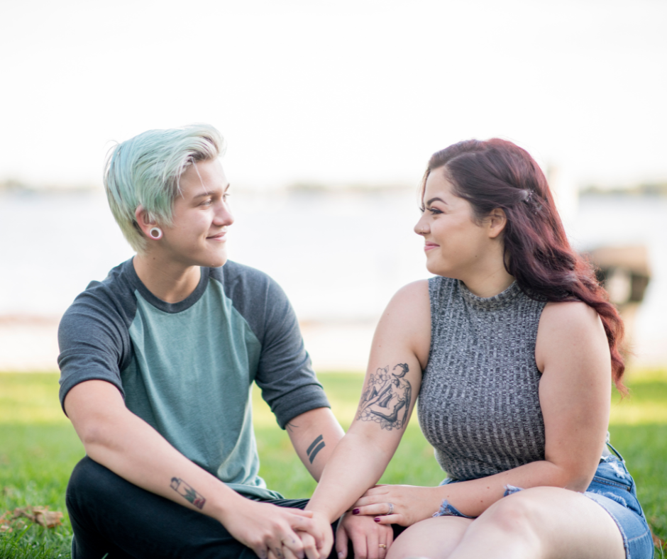 Queer white couple sitting on the grass looking at each other lovingly, holding hands.
