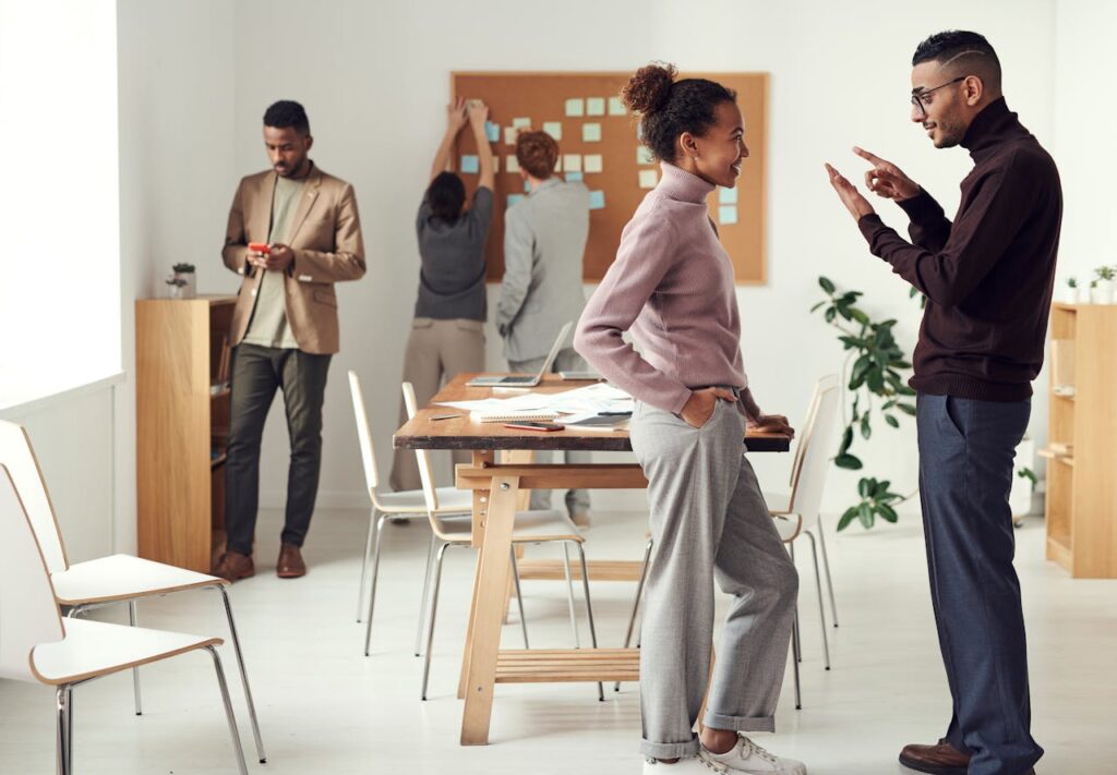 Multiple adults standing around an office room. Two pairs are talking separately, while one individual is alone on their phone. The image highlights different people connecting in different ways.